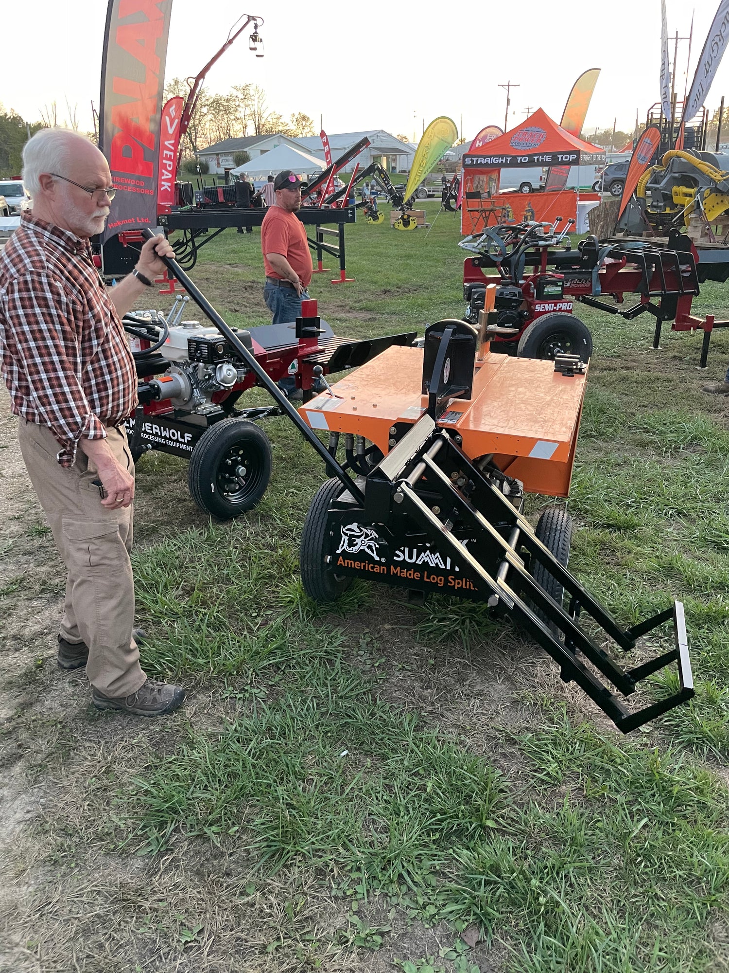 A man is examining a log splitter lift tool at an outdoor event with tents and equipment in the background.